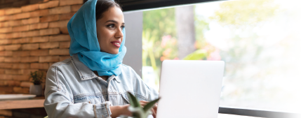A woman wearing a head scarf and typing at a laptop looks out a window.